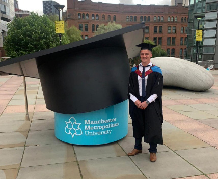Jamie Moore in graduation gown standing next to a large graduation hat