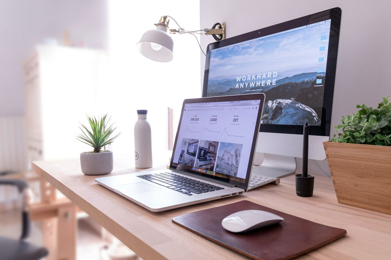A laptop and mouse on a desk infront of a monitor.